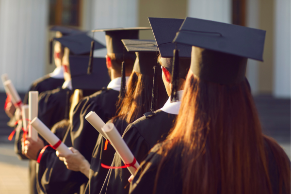 A row of university graduates seen from behind, wearing black caps and gowns and holding rolled diplomas with red ribbons during a commencement ceremony, representing the successful outcome of long-term student retention and academic achievement.